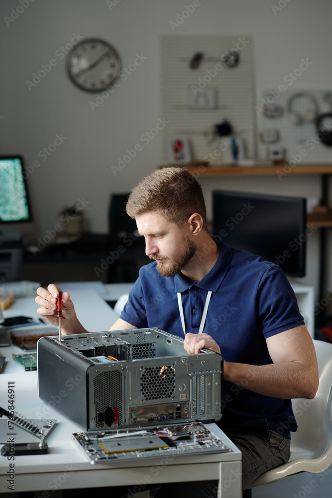 Young technician in uniform sitting by desk in repair service office and using screwdriver while disassembling computer processor - obrazy, fototapety, plakaty 
