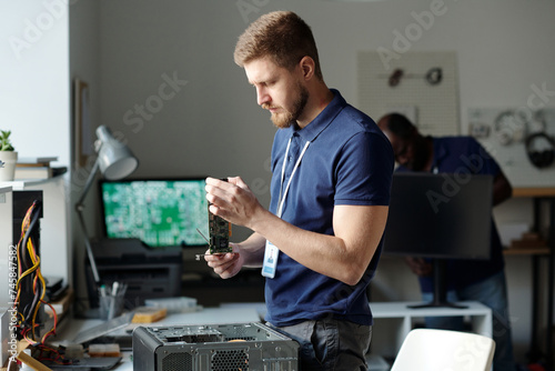 Young male engineer of repair center checking detail of disassembled computer processor while standing by desk with hardware