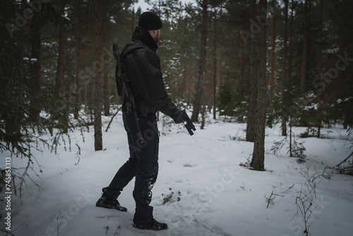 A private military man with an  assault rifle behind his back and a pistol in his hand in a winter forest.