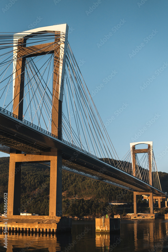 Obraz premium Golden hour illuminates Rande Bridge spanning Vigo Estuary under a clear, azure sky. Reflective waters mirror the bridge, while lush greenery adorns the distant hill under a clear blue sky.