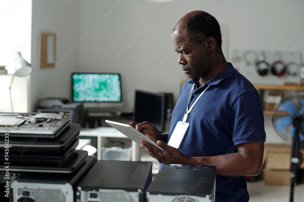 Serious mature African American technician with tablet standing in front of camera by workplace and looking through online manual