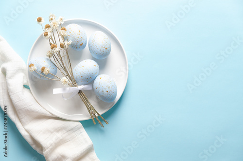 Blue Easter eggs with golden spots and dry flowers in a white plate and a white napkin on a blue background with copy space.