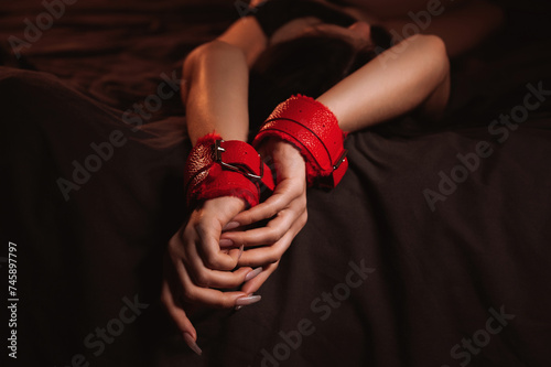 female hands in leather BDSM handcuffs for submission and domination sex on a black sheet in close-up. Submissive woman slave girl lying on the bed in the bedroom
