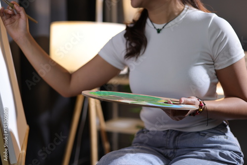 Artist or female painter holding a palette and paintbrush paints and paints her artwork. Young Asian woman drawing in her free time at home.
