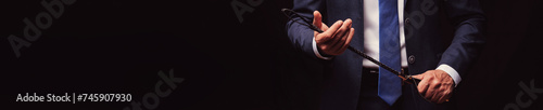 man dominant boss in a suit holds a leather flogger whip in hands for BDSM sex with domination and submission. Wide horizontal banner header with a copy space on black background