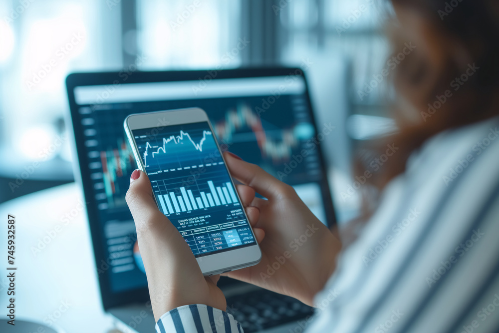 Close-up of investor's hands using smartphone and laptop to study financial statistics and stock market trends.