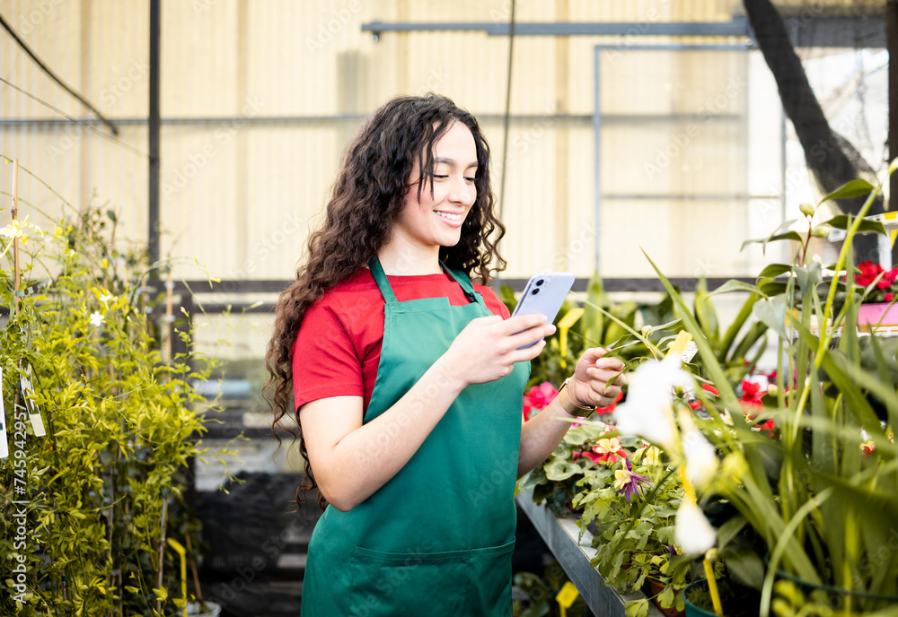 A pretty, Latina girl working inside a plant nursery dressed in a green apron.The young woman takes a photograph of the plants with her mobile phone to upload to social media.Business on social media A pretty, Latina girl working inside a plant nursery dressed in a green apron.The young woman takes a photograph of the plants with her mobile phone to upload to social media.Business on social media