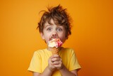 Joyful young boy in a yellow shirt bites a colorful ice cream cone against a vibrant orange backdrop