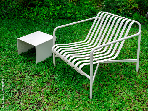 Empty modern minimal design of white empty curved armchair seat and small square side table, iron materials, decoration on green grass background at the summer outdoor backyard garden with nobody.