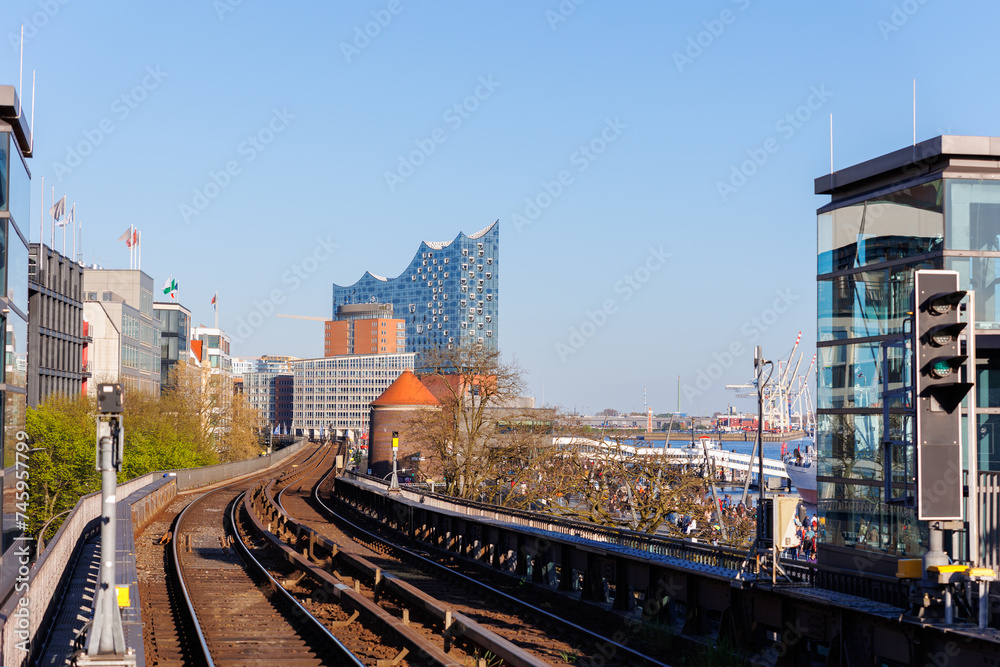 Obraz premium Hamburg Red Unmanned U-bahn Subway train rail Baumwall station on Elevated Track with Hamburger Port Harbor Elbphilharmonie building background blue sky. Hanseatic city commute transport scenic view