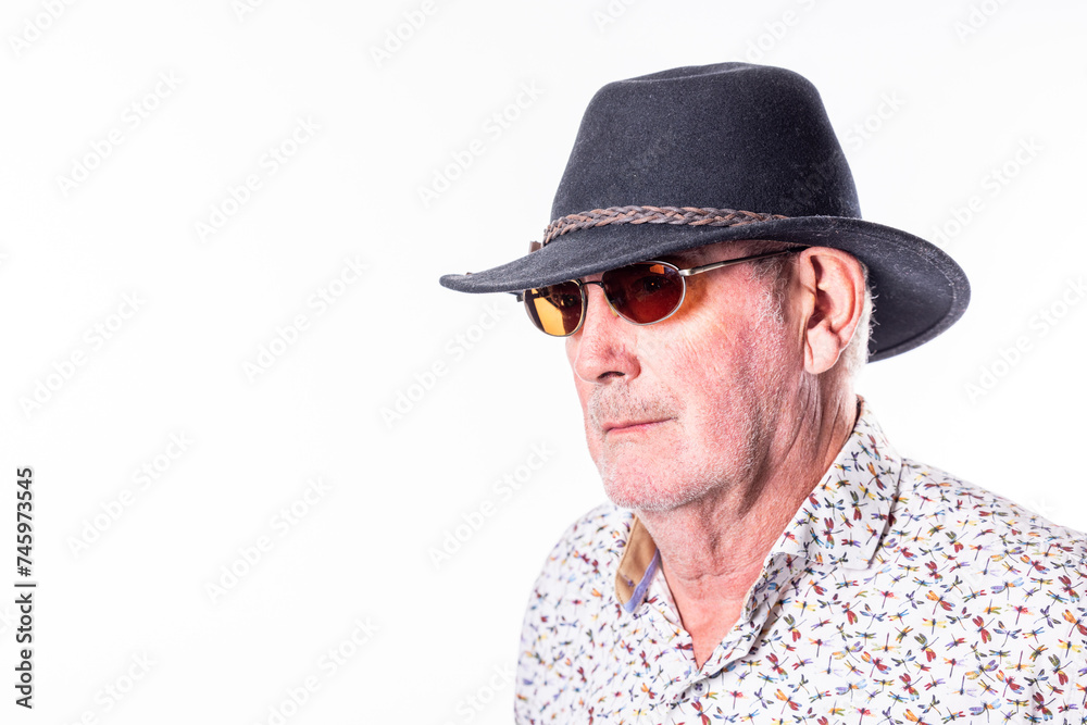 The image depicts an elderly Caucasian man, gazing to the side with a neutral expression. He sports a dark fedora hat and red-tinted sunglasses, which add a touch of style to his persona. The man is The image depicts an elderly Caucasian man, gazing to the side with a neutral expression. He sports a dark fedora hat and red-tinted sunglasses, which add a touch of style to his persona. The man is