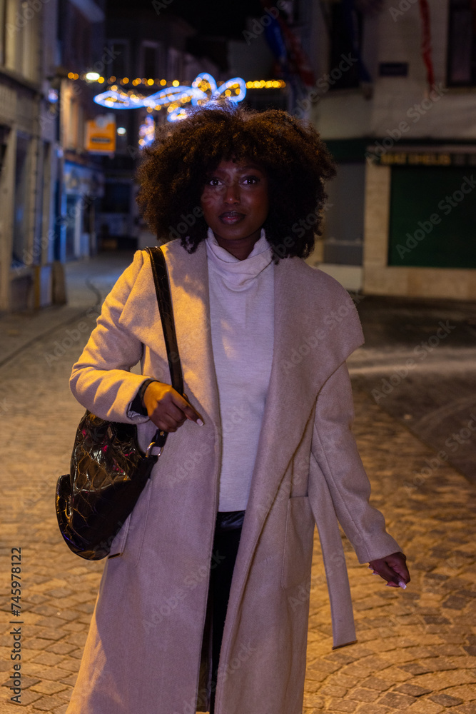 This image portrays a fashionable woman taking a stroll on a cobblestone street at night, illuminated by the ambient glow of city lights and festive decorations. Her confident stance and direct gaze