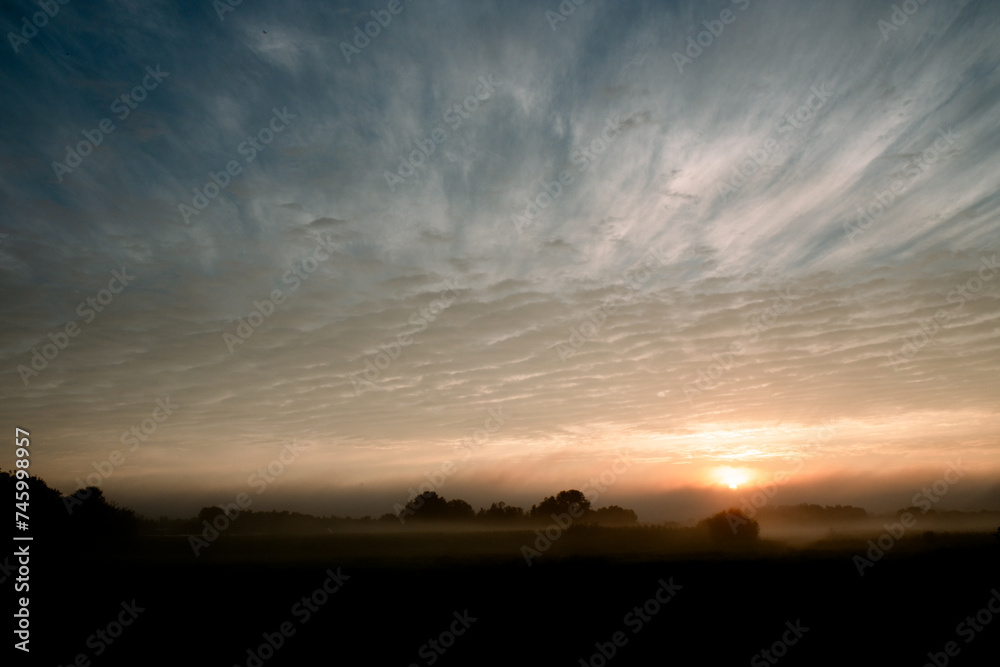 As the day begins, a mystical dawn breaks over fields veiled in mist, with the sun peeking through the low-lying fog, hinting at the landscape's hidden contours. The sky above is a canvas of dramatic