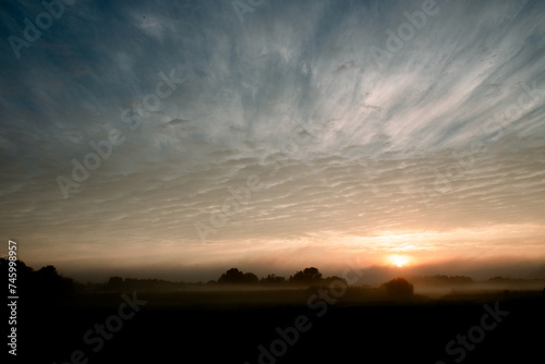 As the day begins  a mystical dawn breaks over fields veiled in mist  with the sun peeking through the low-lying fog  hinting at the landscape s hidden contours. The sky above is a canvas of dramatic