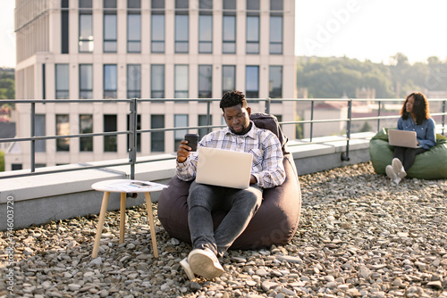 Charming African American man in casual shirt holding cup of hot drink while scrolling webpages on portable computer outdoors, while female colleague working in background. © sofiko14