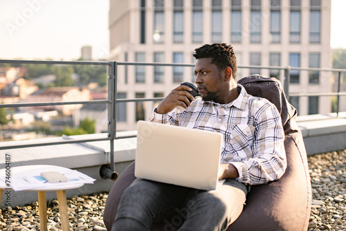 Young man in casual wear carrying out professional activities in open air. Portrait of African male drinking coffee while placing laptop on knees sitting on bean bag chair on roof terrace.