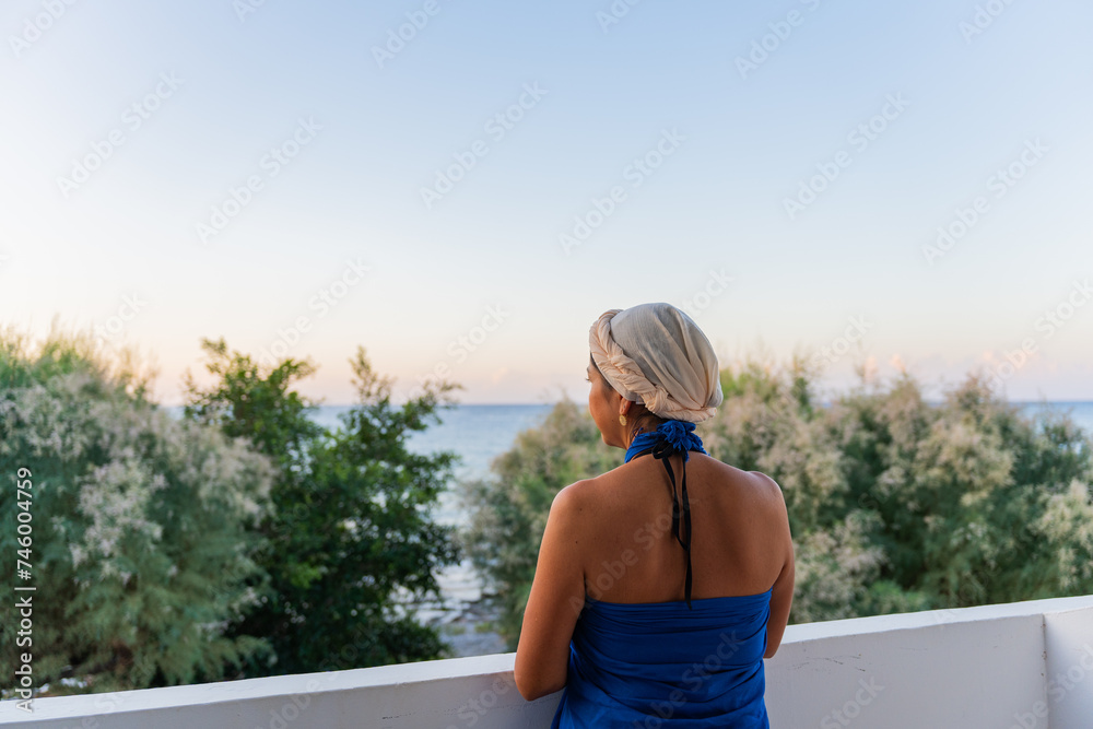 Beautiful woman with blue sarong and Greek outfit on her head enjoying the view from her hotel of the Aegean Sea south of the Greek island of Crete in summer