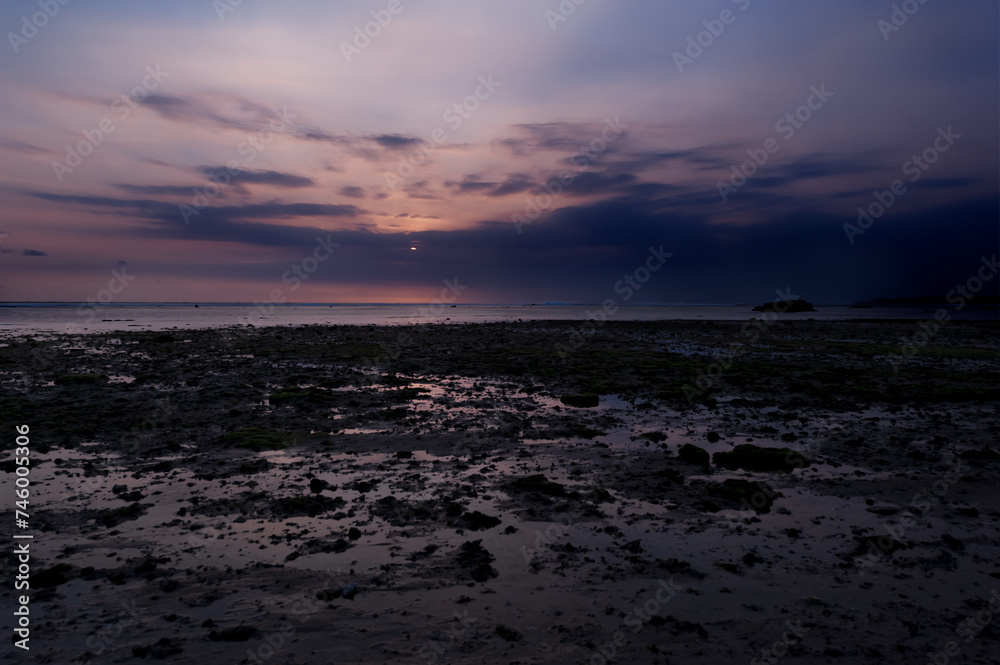 Fototapeta premium Anochecer con sol detras de las nubes y luces rojizas i violetas con algas verdes en Ujung Genteng, Sukabumi, Java, Indonesia