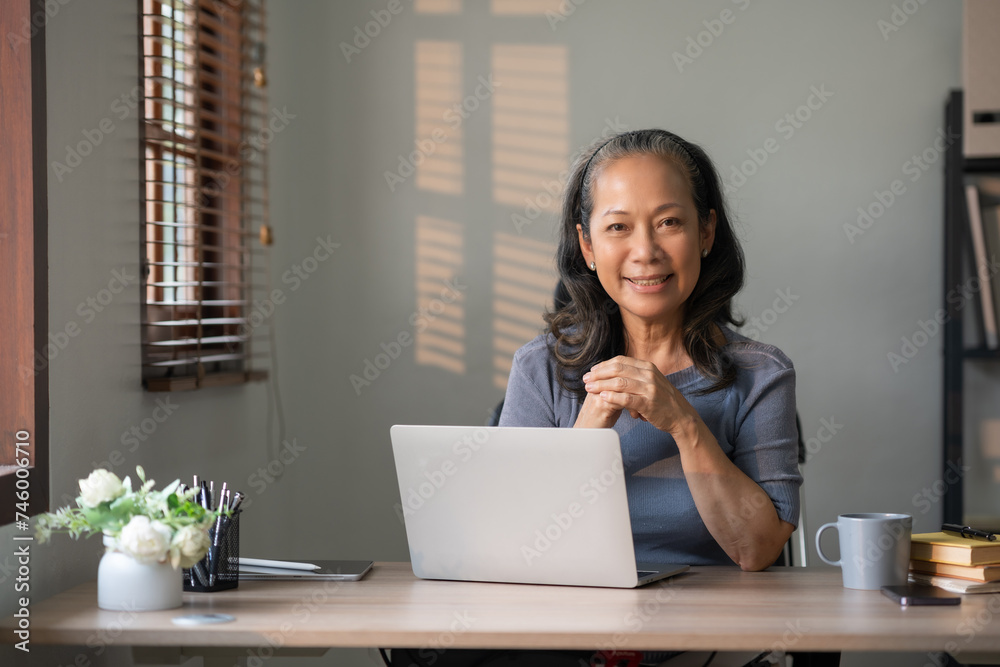 Senior asian woman smiling while working on a laptop at home. Lifelong learning and remote working concept. Senior asian woman smiling while working on a laptop at home. Lifelong learning and remote working concept.