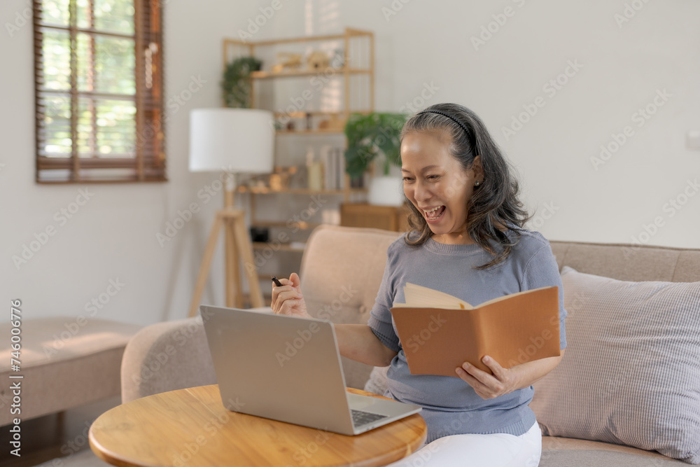 Excited senior Asian woman holding notebook celebrating success with laptop at home Online success and happiness concept. Excited senior Asian woman holding notebook celebrating success with laptop at home Online success and happiness concept.