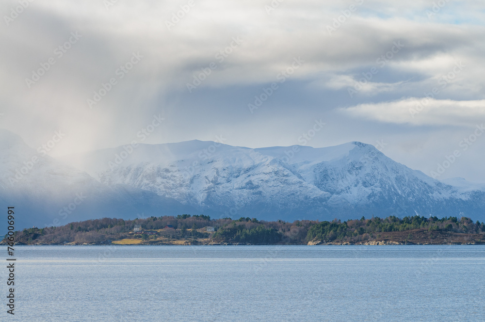 Snowy peaks rise above the tranquil sea with forested shores under a cloudy sky.