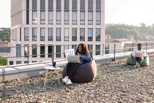 Calm African American businesswoman in denim shirt and headset discussing financial data plan on panoramic terrace with male colleague working on blurred background. © sofiko14