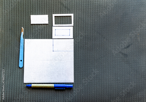 A cardboard paper traced with a felt-tip pen and cut into rectangles hollowed out from the inside. A bistouri knife cutter and a felt pen set on a black board in the background. Overhead view.  photo