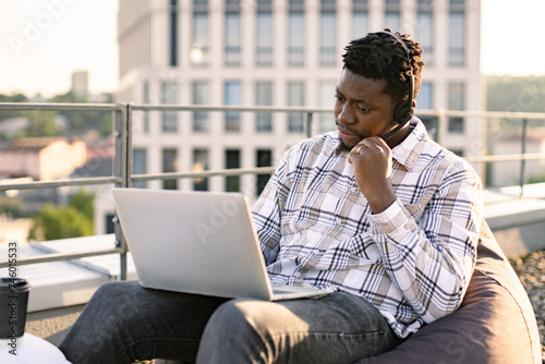 Young African man in casual clothes using portable computer and headset in cozy beanbag on roof terrace. Well-organised employee enjoying open air while staying available during midday. photo