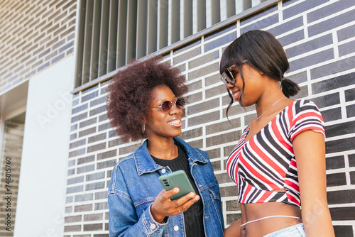 Two sisters share a smile while looking at a smartphone together, their connection evident against a bold, patterned background