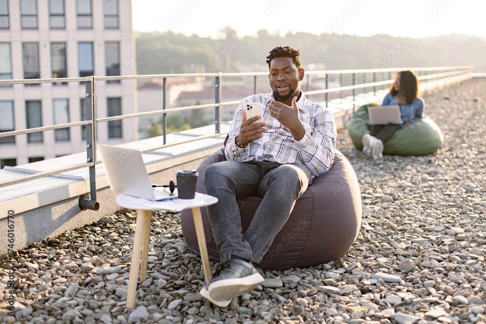 Joyful African man in casual checkered shirt using phone while recording video for her vlog on panoramic terrace, while female colleague working on blurred background. Joyful African man in casual checkered shirt using phone while recording video for her vlog on panoramic terrace, while female colleague working on blurred background.