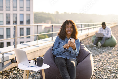 Beautiful African American person with smartphone resting in pouf chair on roof terrace during coffee break on mid day while businessn colleague working on blurred background. photo