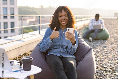 Beautiful African American person with smartphone resting in pouf chair on roof terrace during coffee break on mid day while businessn colleague working on blurred background. photo