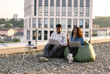 Beautiful young African American couple using laptops while sitting in beanbags at rooftop of office. Two business people working with modern devices sitting on rooftop terrace outside office.