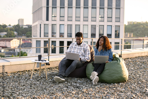 Beautiful young African American couple using laptops while sitting in beanbags at rooftop of office. Two business people working with modern devices sitting on rooftop terrace outside office. © sofiko14