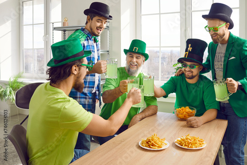 Group of a happy smiling male friends celebrating Saint Patrick s Day at home. Joyful men in green hat and glasses having fun at the party drinking beer with french fries and chips.