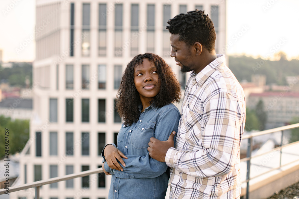 Young African American couple woman and man hugging on rooftop of modern building and looking away. Concept of love relationships and outdoors activity.