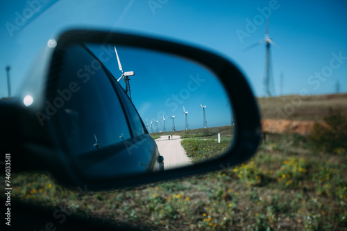 A car mirror that reflects the road and white windmills. Wind turbines alternative energy generation