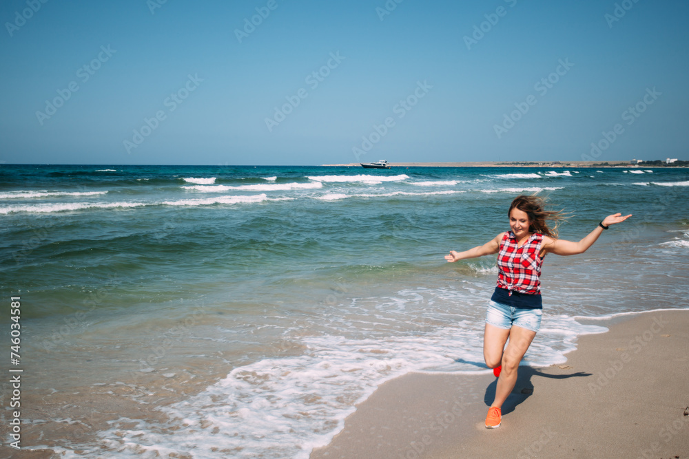 A girl in denim shorts and a red plaid shirt runs along the ocean on the snow-white sand. The woman is happy and has her arms outstretched. The sea waves are coming in, the sea is turquoise