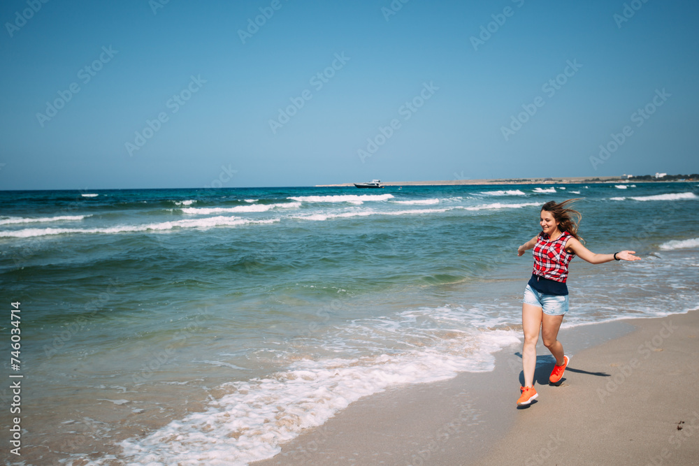 A girl in denim shorts and a red plaid shirt runs along the ocean on the snow-white sand. The woman is happy and has her arms outstretched. The sea waves are coming in, the sea is turquoise