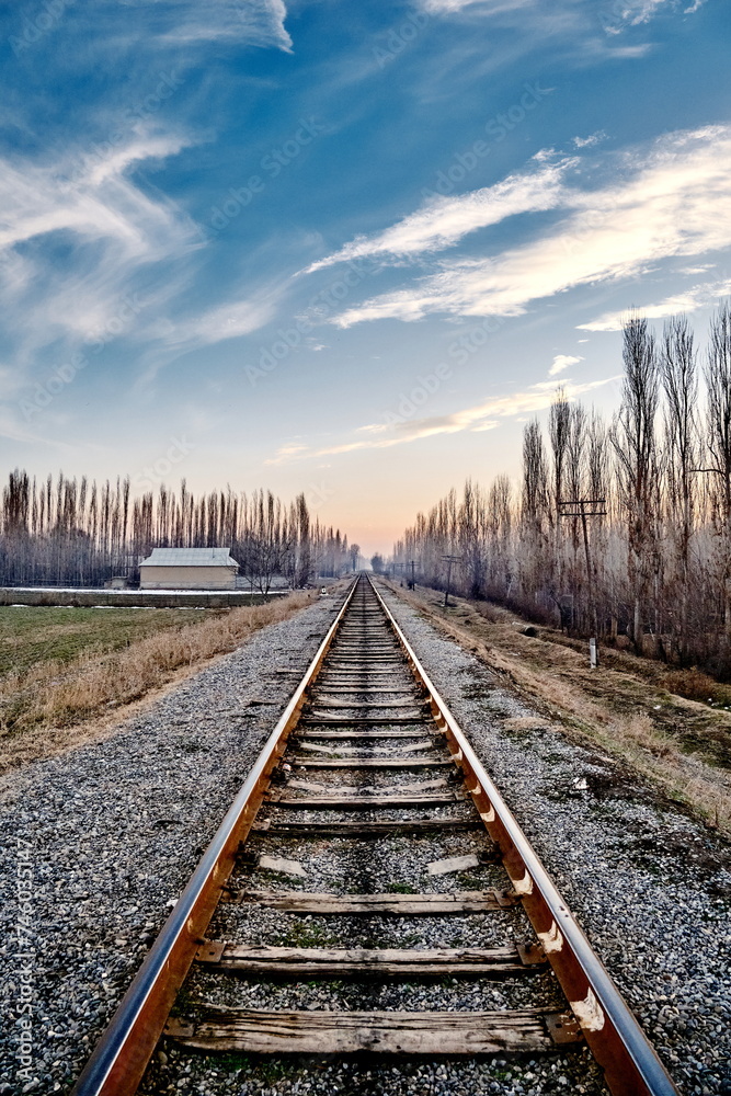 Obraz premium A railroad disappearing into the distance amidst fields in rural scenery, accompanied by old wooden electrical poles running alongside, against the backdrop of a sunset sky and distant light mist.