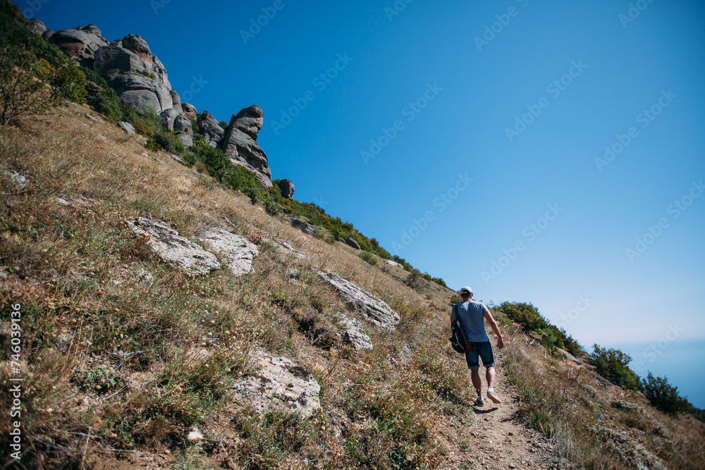 A male tourist is climbing up a mountain trail. A man with a backpack is doing mountain hiking. Hiking in the mountains in summer