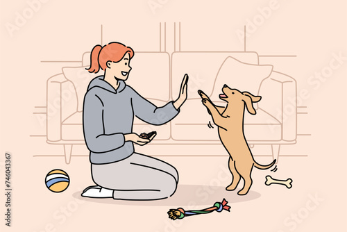 Woman trains dog, sitting on floor near sofa and giving food to puppy after following command. Smart dog stands on two legs, fulfilling requirements of female trainer who enjoys communicating with pet