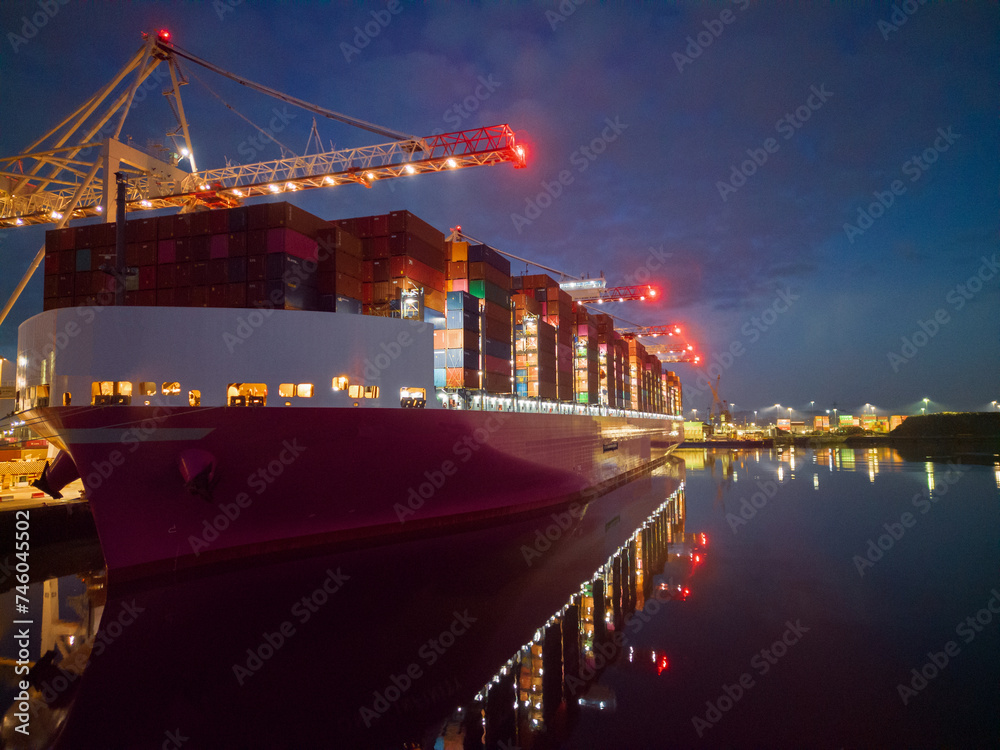Pink container ship at night at Southampton Docks, UK. Aerial view of the bow and side view of the vessel loaded with shipping containers. Calm sea with ship and port crane lights reflection.