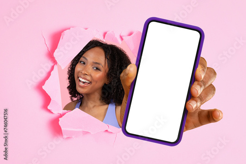 Cheerful woman peeking through a torn pink paper wall presenting a smartphone with a blank white screen © Prostock-studio