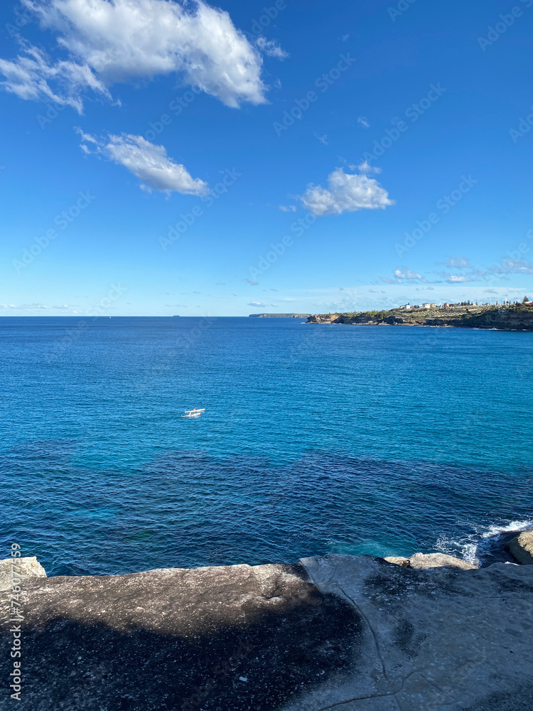 Ocean and sky. Bay and rocky coast with turquoise waves breaking on the shore. View of the town and people who row in the distance. Australian landscape and shoreline.