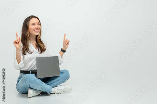 Joyful young woman sitting cross-legged on the floor, holding a laptop with one hand © Prostock-studio