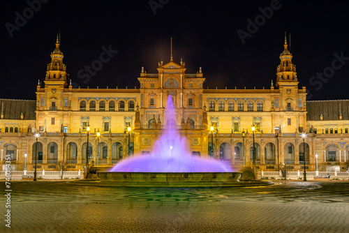 Vista nocturna de la Plaza de España de Sevilla al anochecer, con la fuente central iluminada de morado