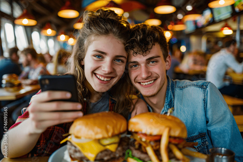 A happy couple taking selfies on a smartphone in a burger pub restaurant  surrounded by a lively atmosphere of young people relaxing for lunch in a cafe bar.