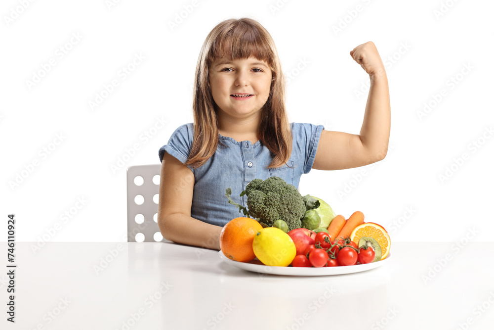 Happy little girl sitting with a plate of fruits and vegetables on a table and showing muscle Happy little girl sitting with a plate of fruits and vegetables on a table and showing muscle