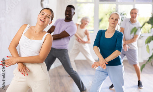 Young girl coach oversees mentoring group of elderly clients in fitness studio. Multiracial group of different ages practice repeating dance movements